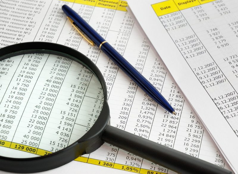 a magnifying glass atop financial papers with a pen, some yellow highlights appear in the headers of the pages